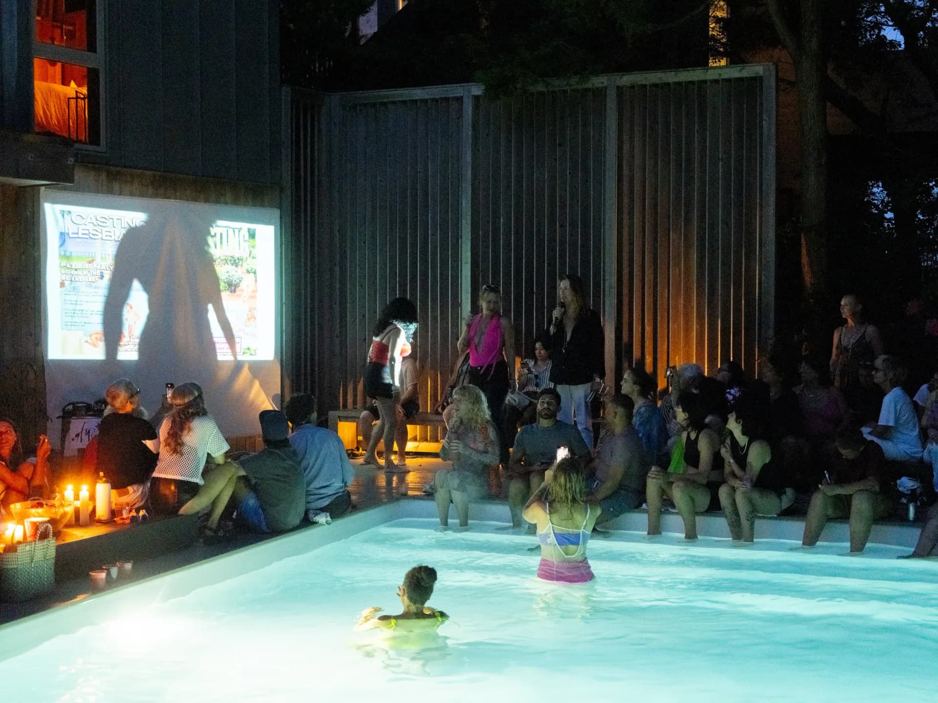 Photograph of AMR Art Dumbo's interior space at night: illuminated pool with person swimming, corrugated metal walls, gathered visitors, projection screen, and ambient lighting.