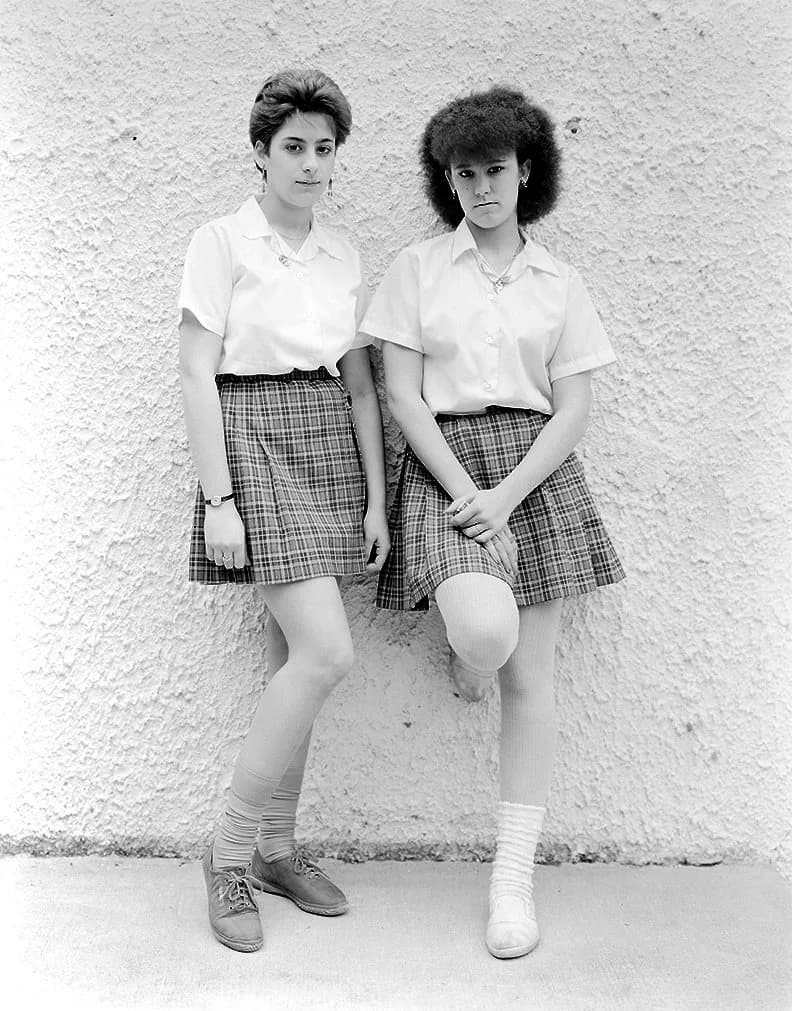 Black-and-white photograph of two women standing against white wall, wearing matching white shirts and plaid skirts with period-appropriate 1980s styling.