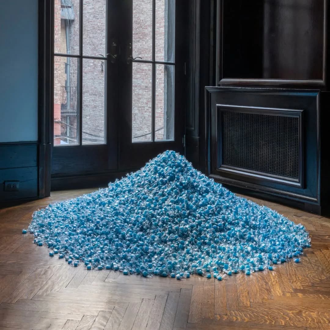 A large conical pile of turquoise and blue granular material on a wooden parquet floor in a gallery space with black-framed windows and doors.
