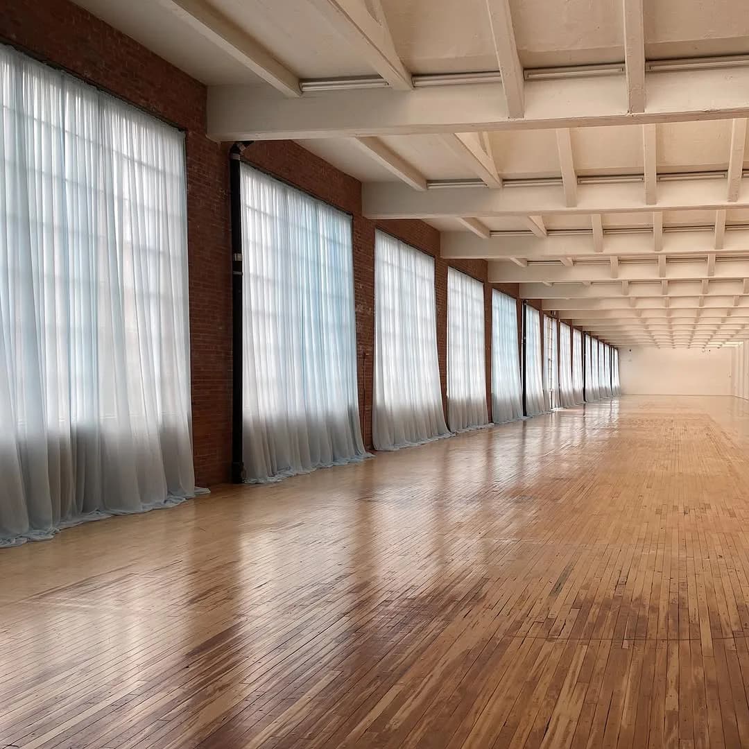 Long gallery corridor with exposed brick walls, white coffered ceiling, wooden herringbone flooring, and sheer curtains covering tall windows along the sides.