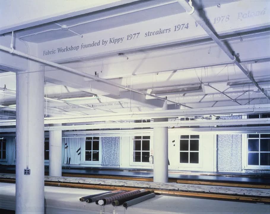 Industrial warehouse interior with white columns and ceiling beams, text reading 'Fabric Workshop founded by Kippy 1977 streakers 1974', and a dark sculptural form on the floor.