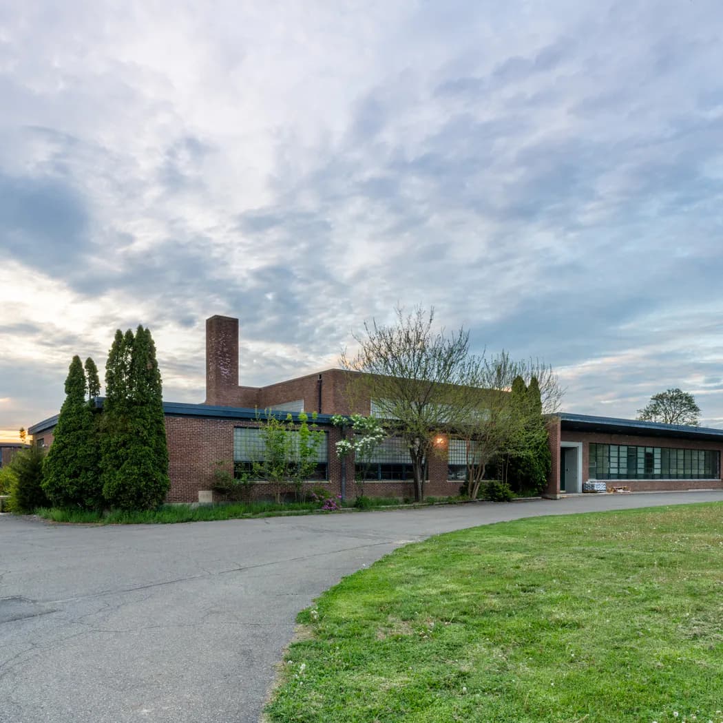 Exterior of Andrew Kreps Gallery, a modernist brick building with tall windows and a tall chimney, surrounded by manicured lawn and cloudy sky.