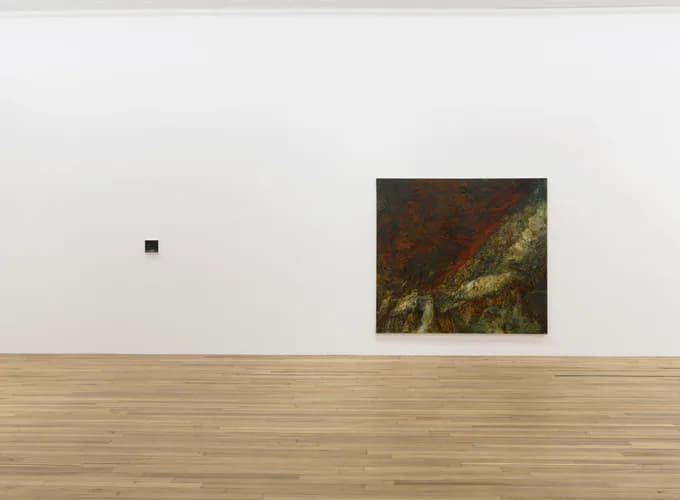 Gallery interior with white walls and wooden floor, displaying a small dark artwork on far left wall and a larger dark painting on right wall.