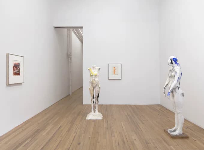 Gallery interior with wooden floors and white walls, featuring small framed artworks, two figurative sculptures, and an open doorway.