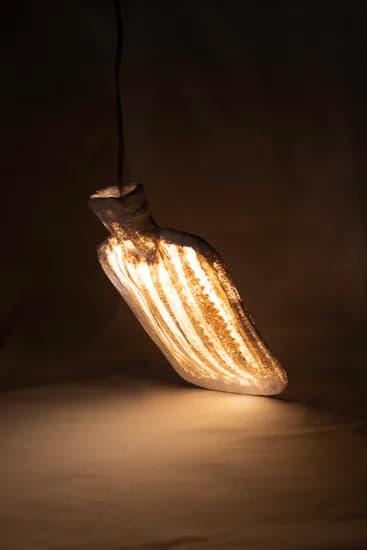 Illuminated sculptural form resembling a curved leaf or organic shape with vertical ribbed details, casting warm golden light against a dark background.