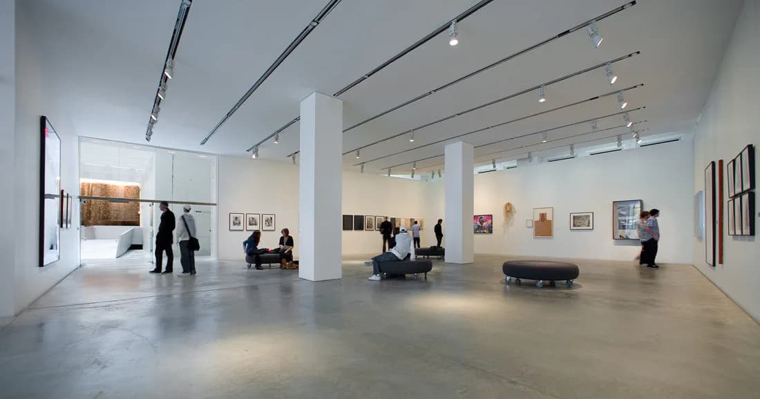 Contemporary gallery interior with high ceilings, polished concrete floors, white walls with artwork installations, and visitors.