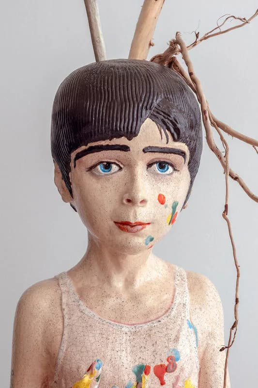 Wooden sculptural bust of a figure with short dark hair, blue eyes, colorful marks on face and neck, and wooden branches extending from the top.