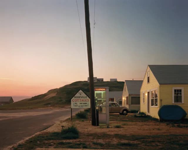 Color photograph of a cottage with yellow siding at dusk, a utility pole, and a hillside in warm sunset light.