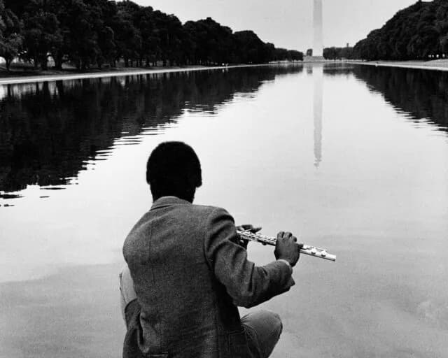 Black and white photograph of a person from behind holding a flute, standing by a reflecting pool with a distant monument visible.