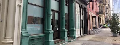 Storefront exterior in Tribeca with green-painted facade, large windows, and adjacent brick buildings on a quiet street.