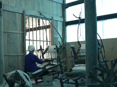 Person seated at machinery inside an industrial structure with metal framework and large barred window opening to exterior.