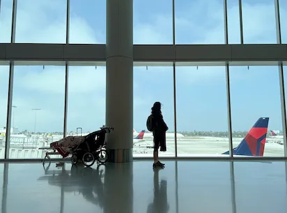 Person with stroller standing at airport terminal window overlooking runways and aircraft in daylight.