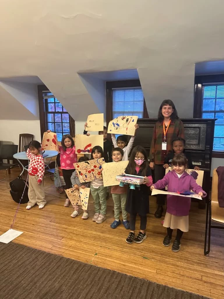 Group of children holding large colorful artwork pieces in an angled-ceiling studio space with windows and piano.