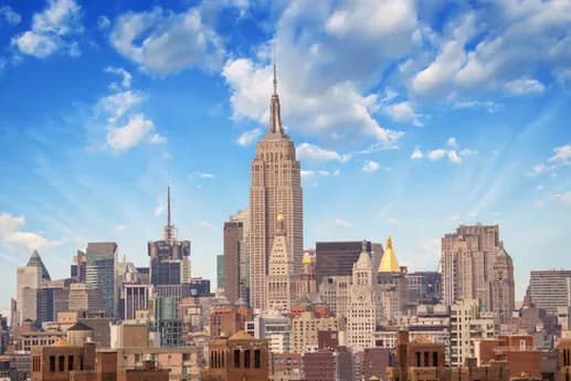 Manhattan skyline photograph dominated by the Empire State Building's golden Art Deco spire, surrounded by historic and modern skyscrapers under blue sky with white clouds.