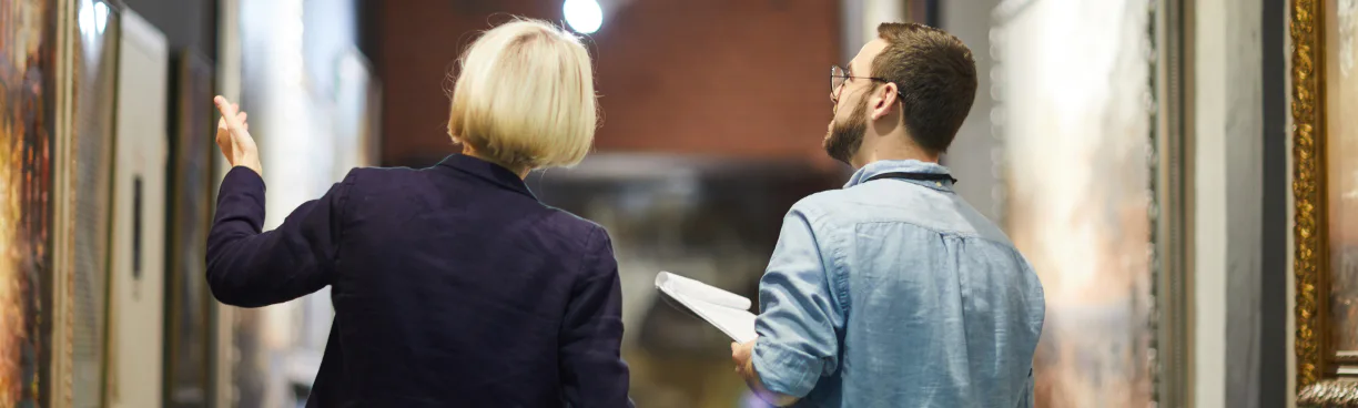 A blonde woman and a bearded man viewed from behind examine framed paintings hanging in a dimly lit Art Peritus gallery hallway