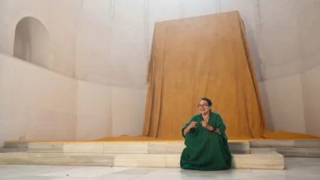 Woman in a green robe sits on marble steps before a large draped golden fabric installation inside a domed memorial-like interior, part of an Art21 feature.