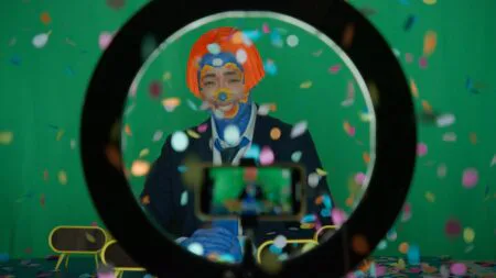 Person in orange wig and face paint sits before a green screen, viewed through a ring light with confetti falling and a phone recording in the foreground, from an Art21 video.