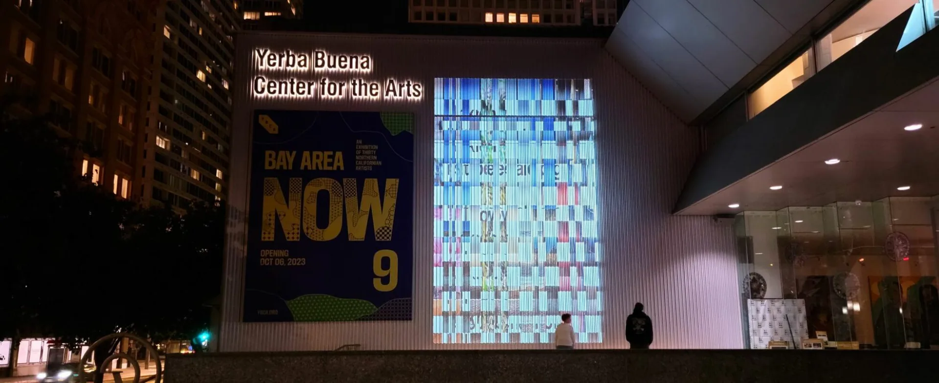 Nighttime exterior of the Yerba Buena Center for the Arts with an illuminated 'Bay Area Now 9' banner and a large lit grid installation on the facade