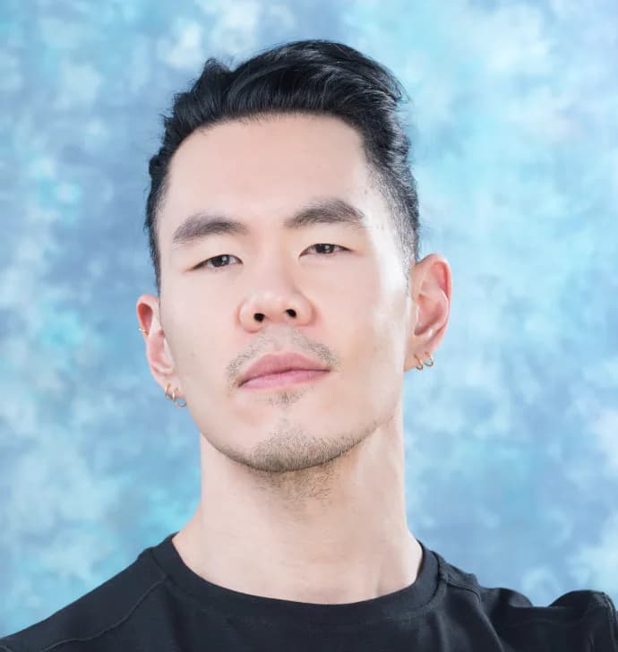 Close-up studio portrait of a man with slicked-back dark hair wearing a black shirt against a mottled blue background