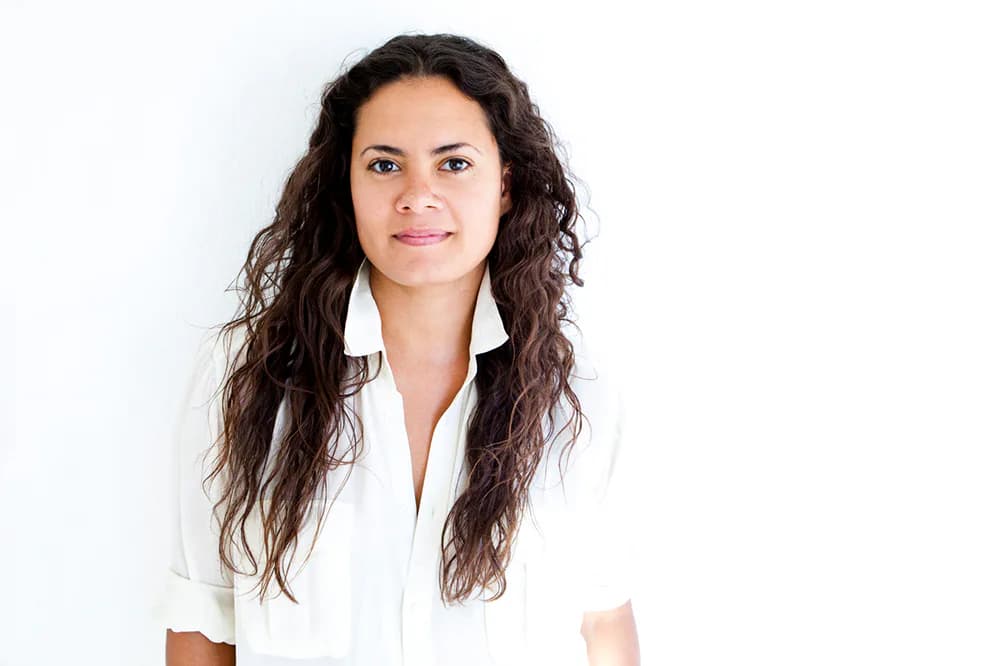 Woman with long curly brown hair wearing a white blouse, smiling softly against a plain white background