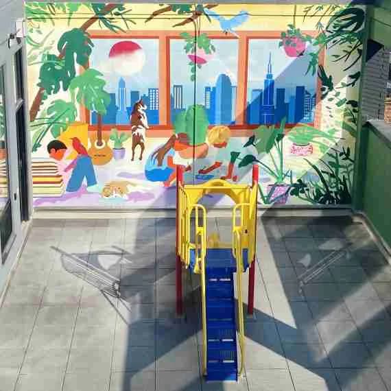 Colorful window installation with painted figures, cityscape, tropical foliage, and playful composition in vibrant blues, greens, oranges, and pinks.