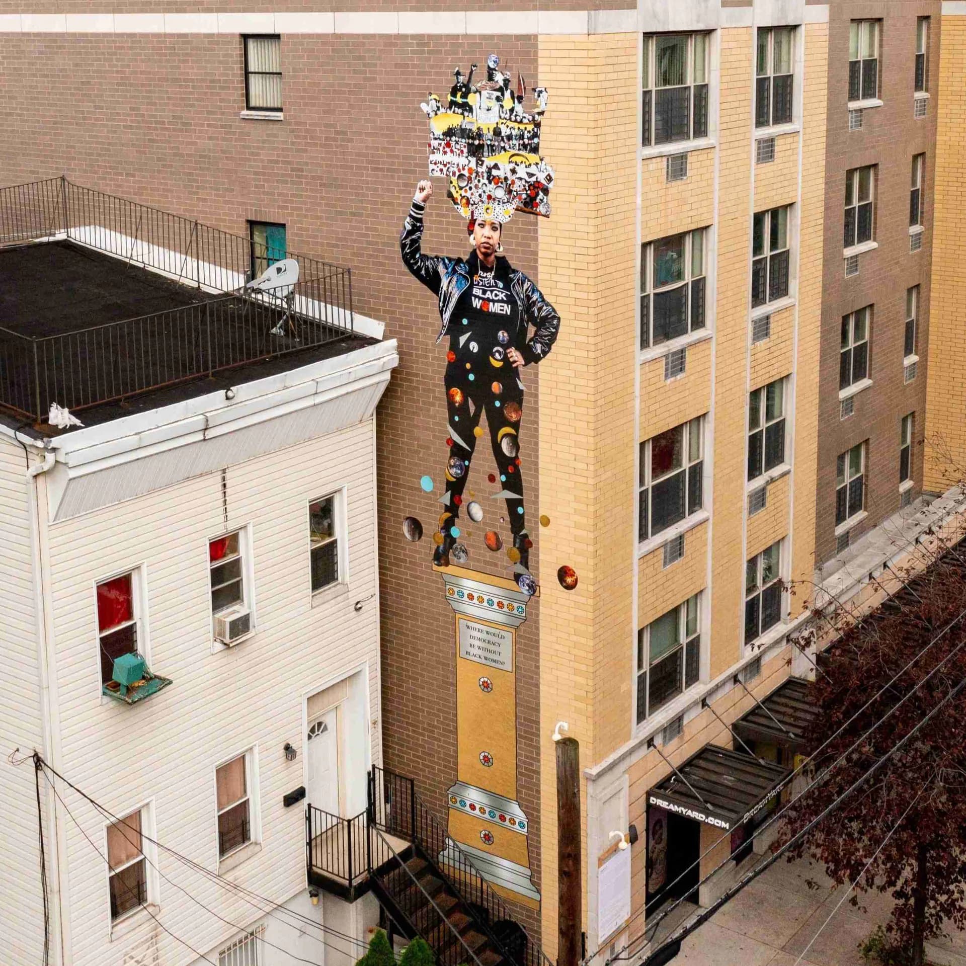 Tall sculptural figure in navy with yellow accents reaching upward on exterior brick building wall, with floating objects above.