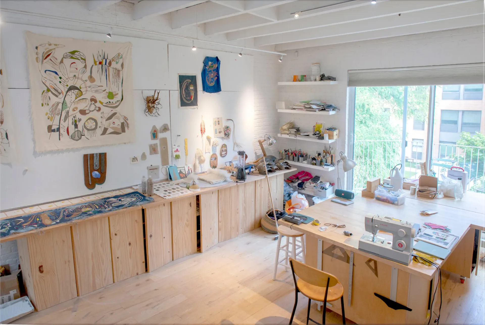 Bright studio interior with wooden cabinetry, work tables, wall-mounted artwork, shelving, and large windows overlooking urban landscape.