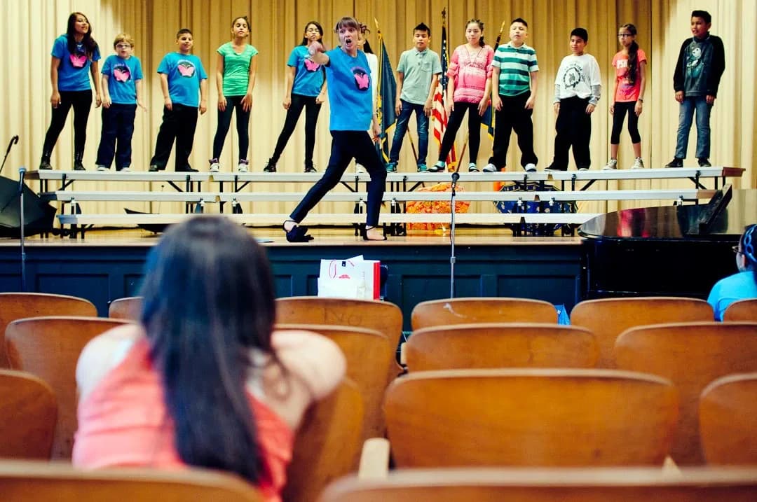 Young performers in colorful blue, pink, and green shirts stand on an auditorium stage above wooden seats, with one person mid-jump on a balance beam.