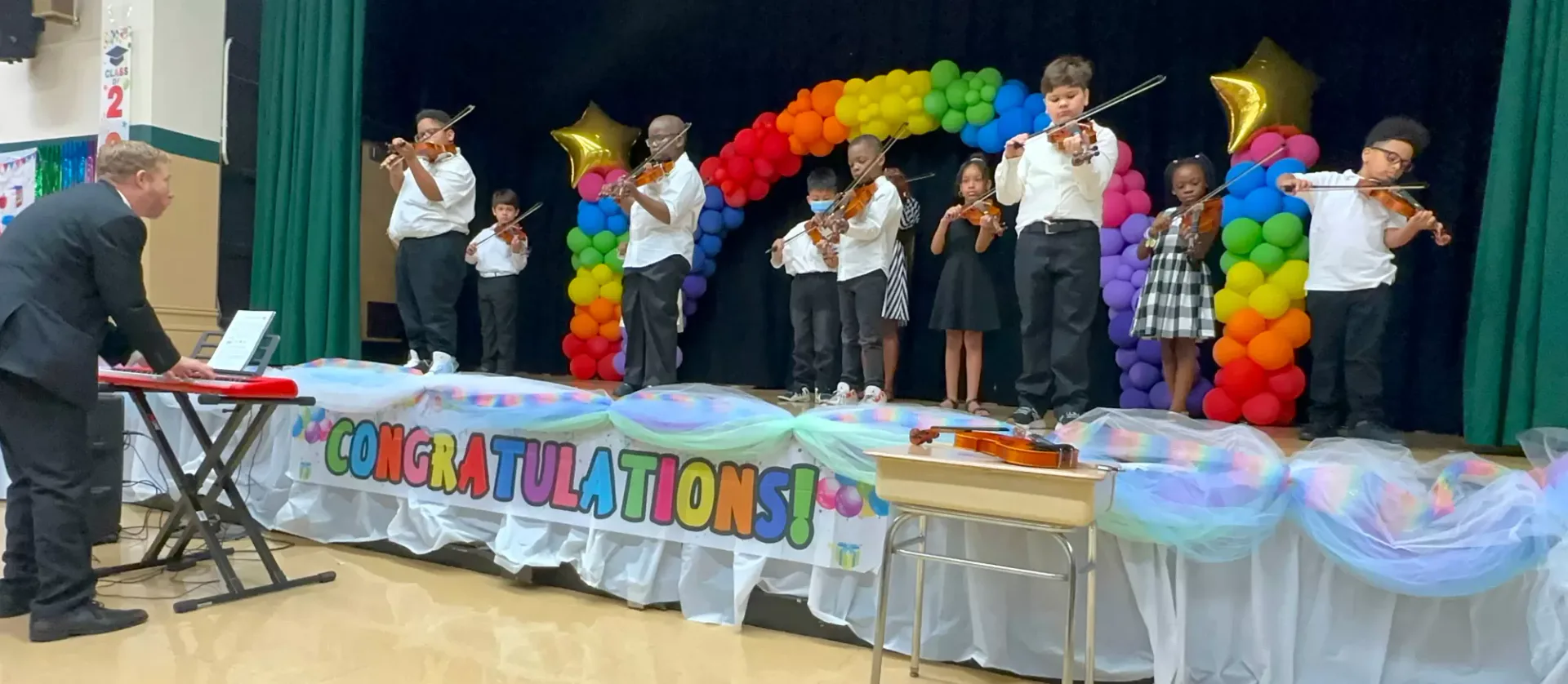 School gymnasium stage with children standing before a colorful balloon arch and congratulations banner during an Arts Horizons celebration event.