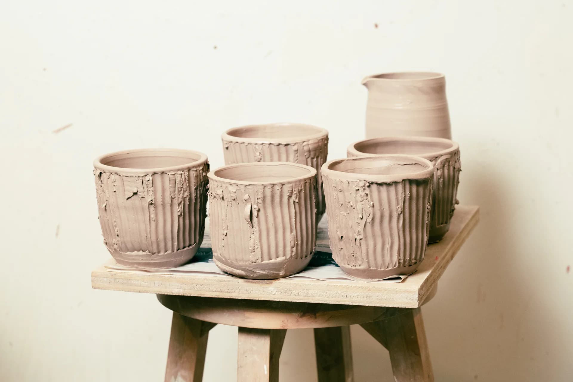 Five clay vessels with vertical ribbed texture displayed on white platform and wooden stool against neutral background.