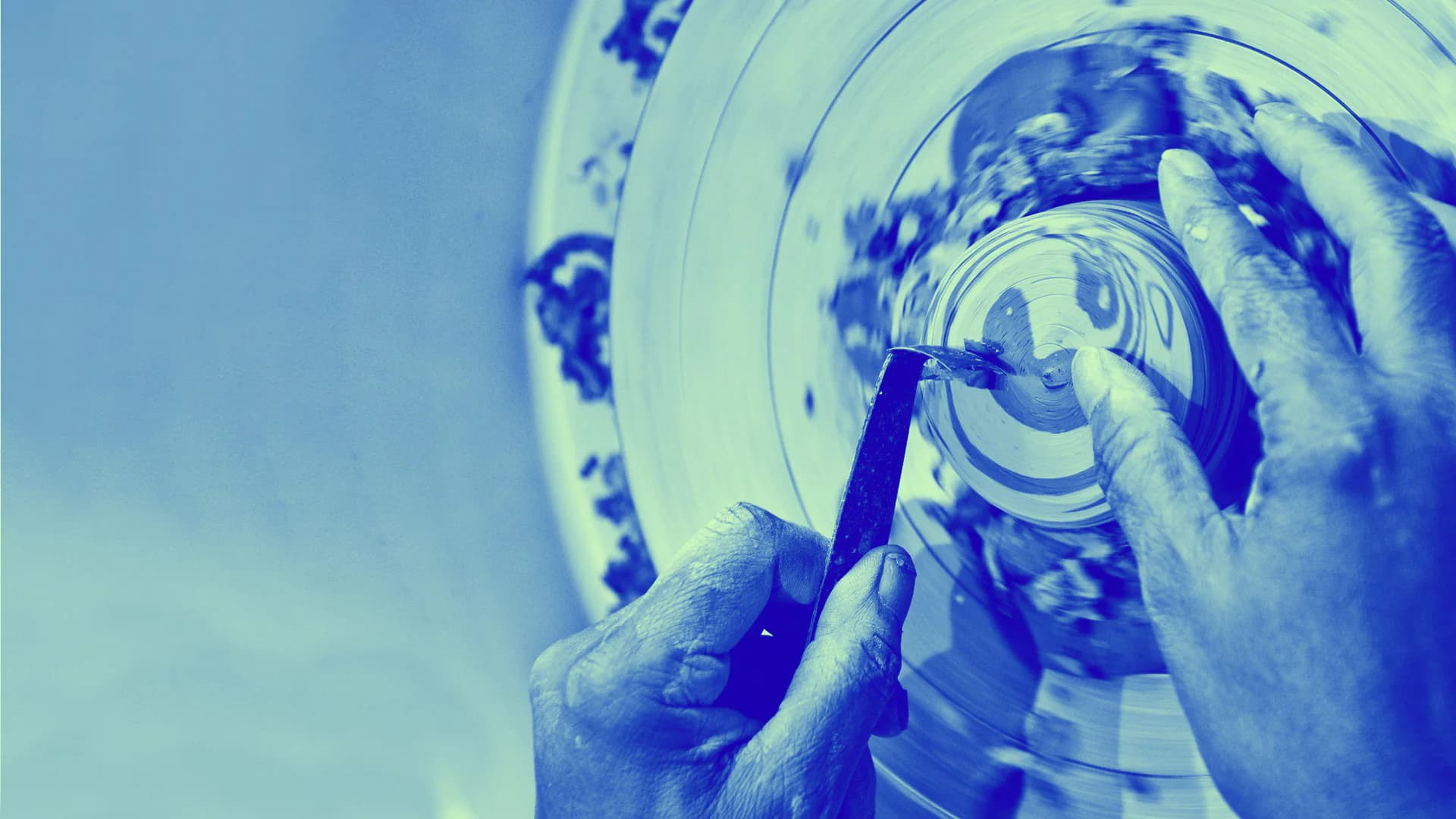 Close-up blue-toned photograph of hands working on pottery wheel with clay vessel in progress.