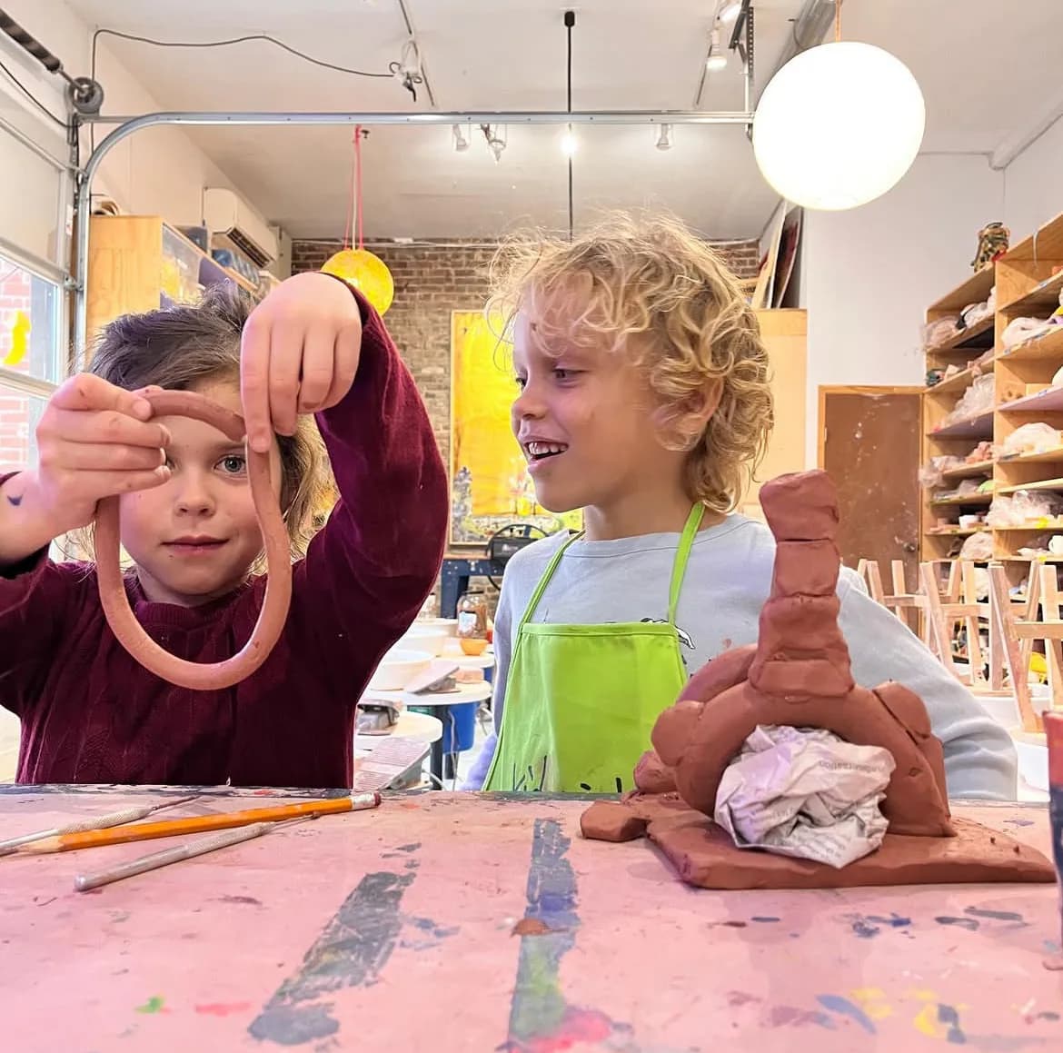 Two children in workshop space holding and displaying clay sculptural pieces they created, with pink work surface and shelving visible.