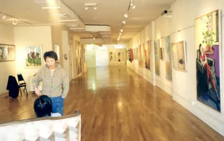 Gallery interior at Asian American Women Artists Alliance with polished wood floors, white walls displaying framed artworks, track lighting, and a woman sitting in the foreground.