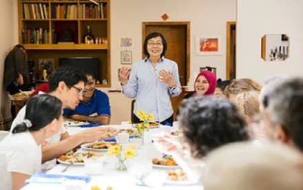 Gallery workshop or community event with diverse participants seated around a table with food, a woman standing and speaking, and bookshelves visible in background.