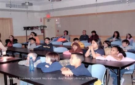 Gallery or library workshop space with children and adults engaged in a shadow puppet activity at long tables with an instructor leading the session.