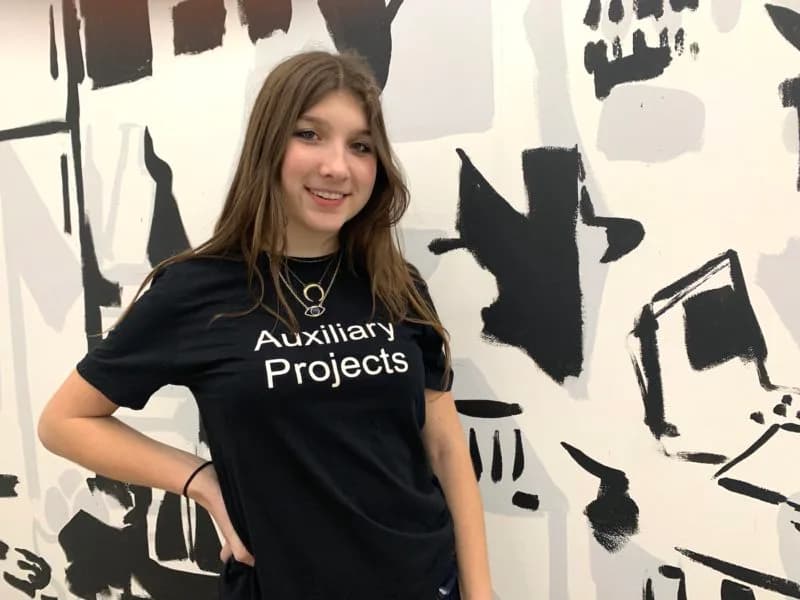 Woman wearing black t-shirt reading 'Auxiliary Projects' against abstract black and white wall art.