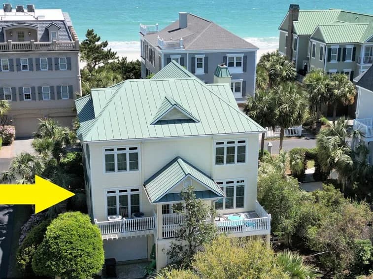 Aerial photograph of a two-story white house with green metal roof and white railings, surrounded by landscaping and neighboring coastal homes under blue sky.