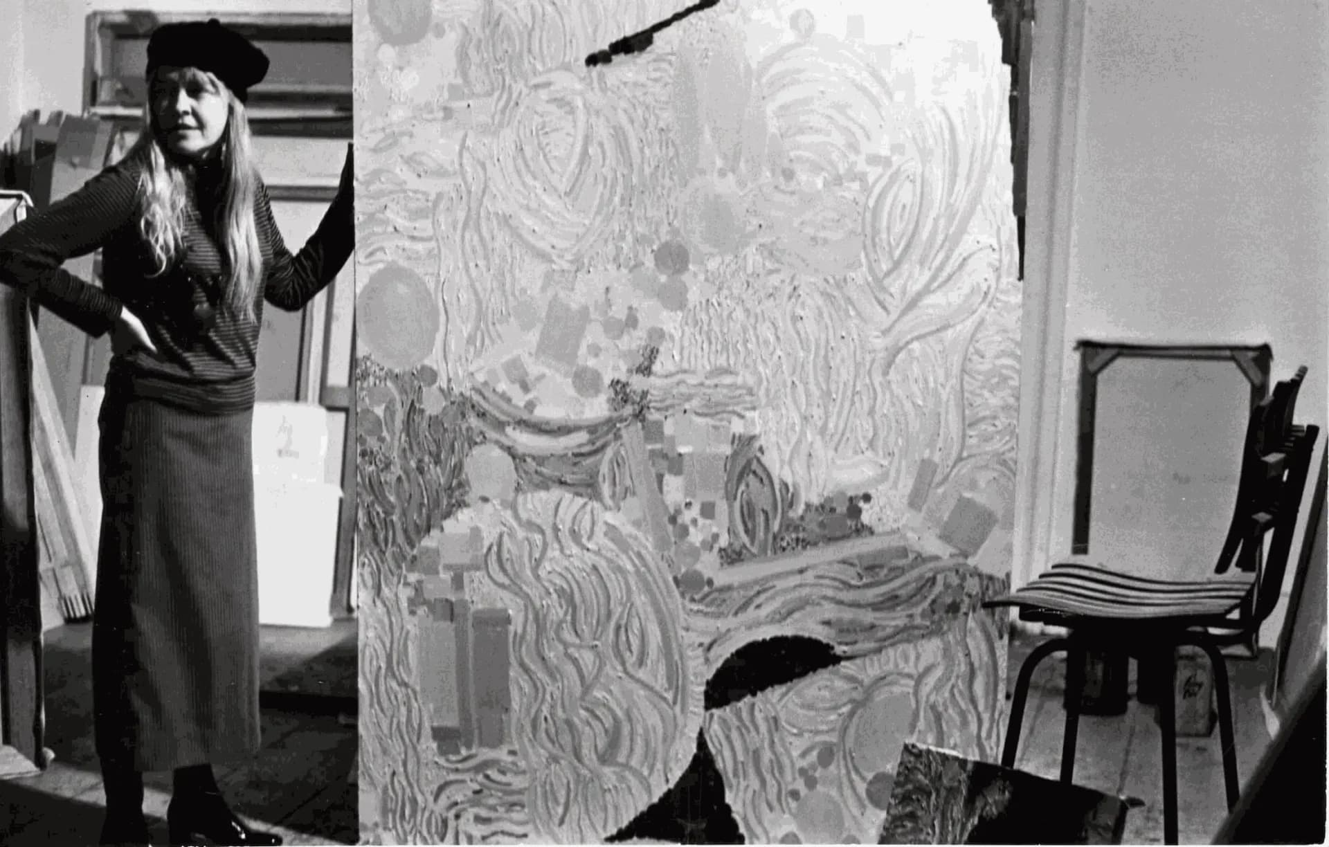 Black and white photograph of a person with long hair standing next to a large drawing or artwork with swirling, expressive line work.