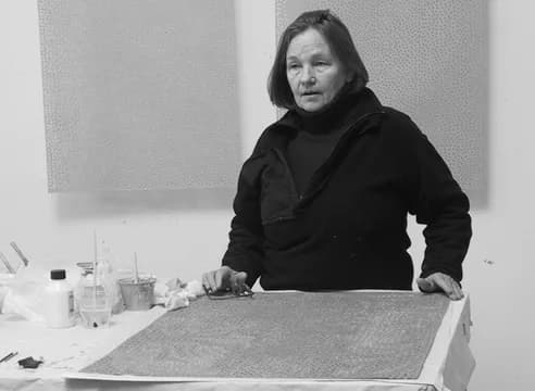 Black and white photograph of a woman with dark hair wearing a black jacket, standing at a work table in a gallery space.