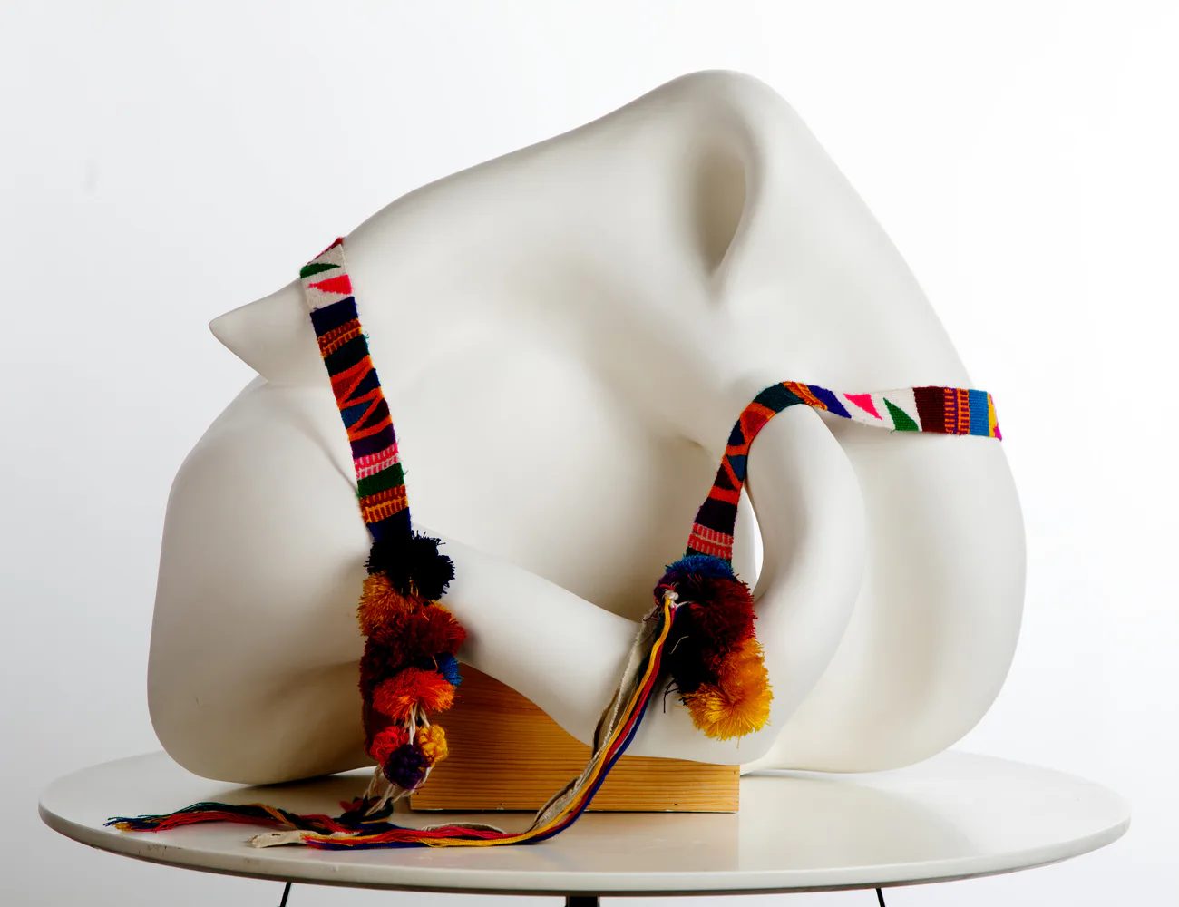 Abstract mixed-media sculpture: cream-colored curved form with colorful woven textile straps and wooden element, displayed on white platform.