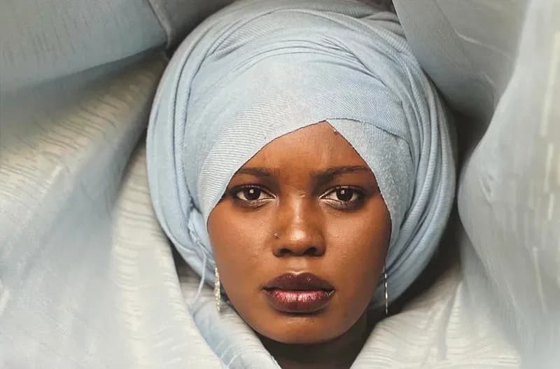 Portrait photograph of a person with light-wrapped headwrap against white bedding, direct gaze at camera.