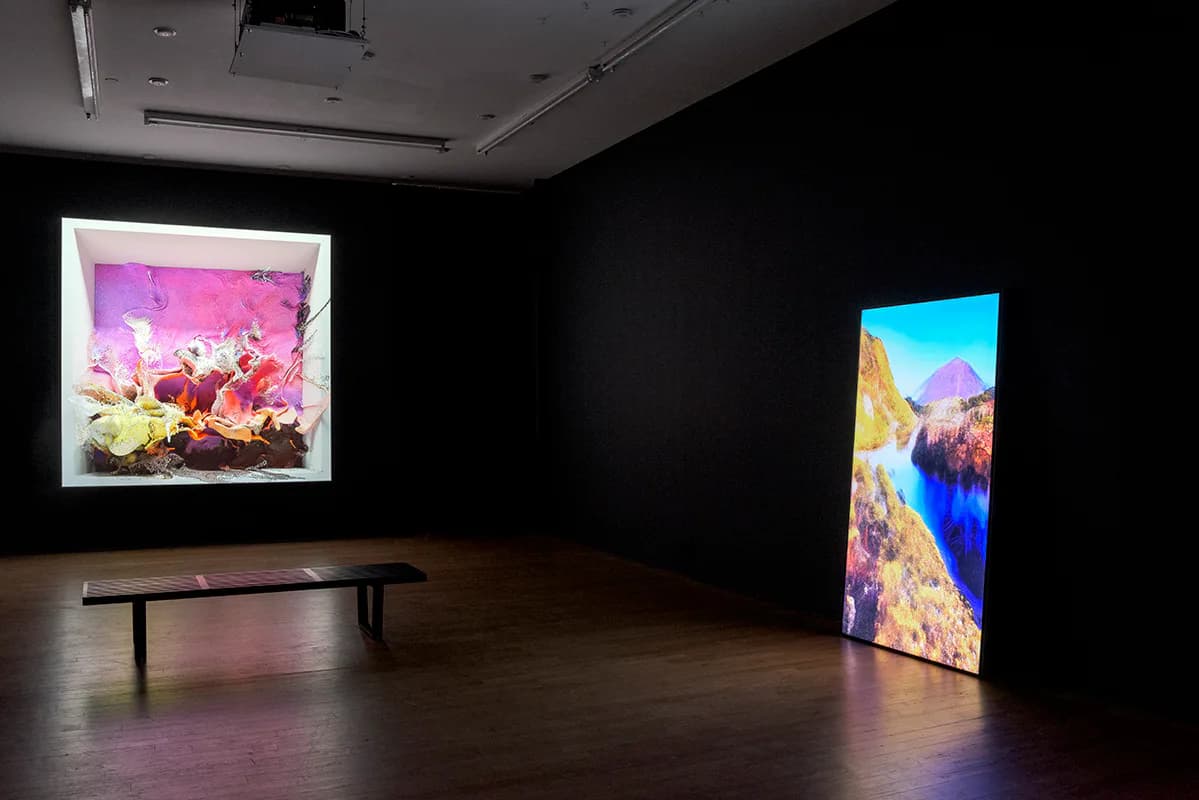 Gallery interior with dark walls featuring two illuminated digital artworks: a square screen displaying pink and yellow floral imagery on the left, and a tall vertical screen showing a colorful landscape on the right, with a wooden bench in the center.