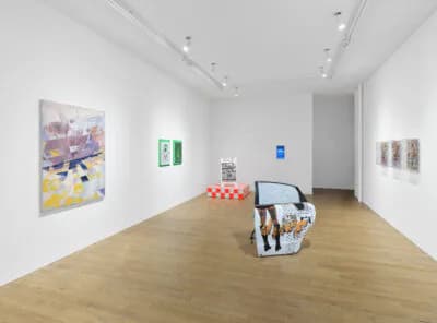 Gallery interior with white walls and wooden floor displaying mixed-media artworks, including colorful abstract paintings and sculptural objects, with bright overhead track lighting.