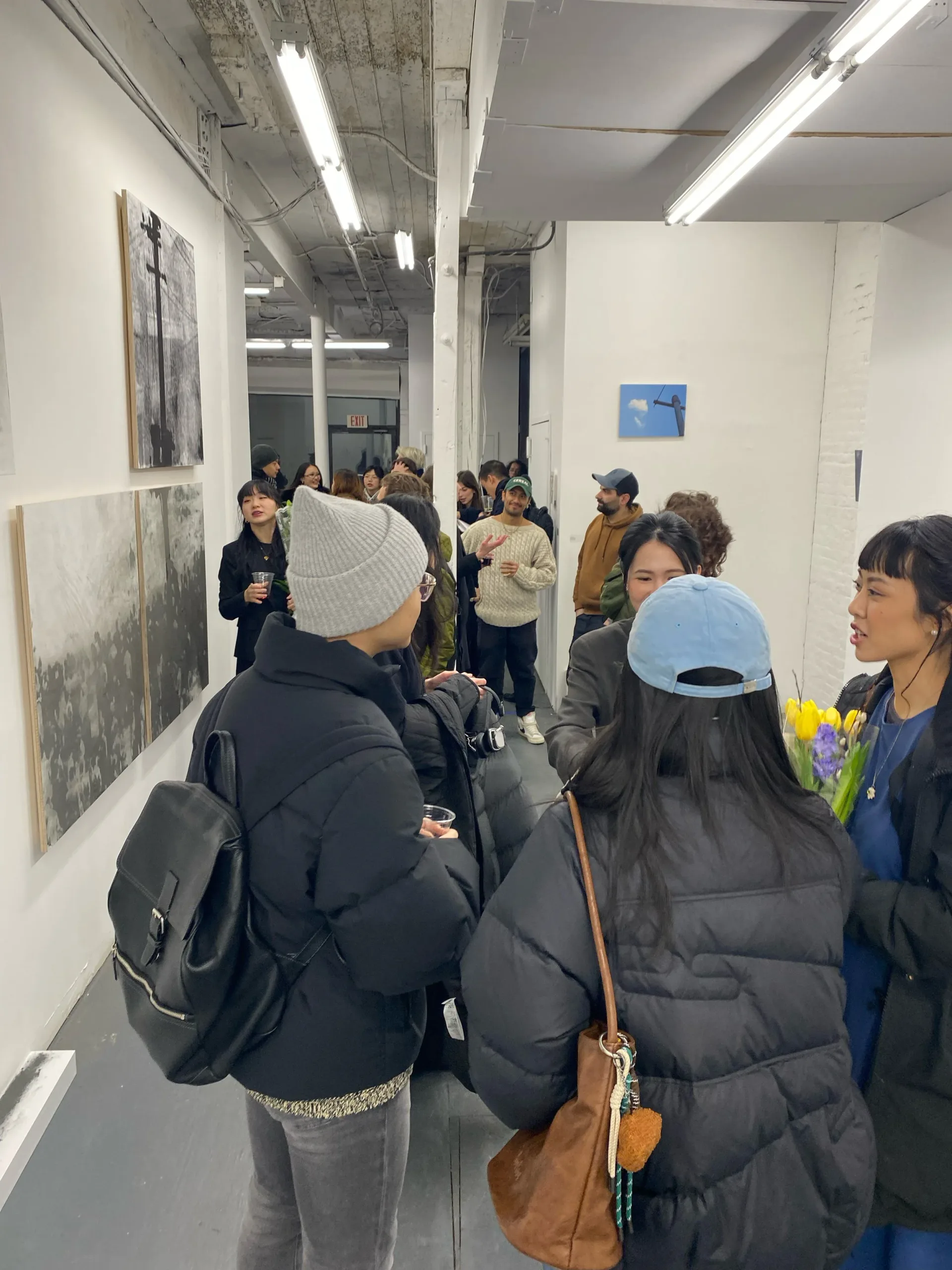 Gallery corridor with visitors viewing artwork on white walls under fluorescent lighting; gray and white abstract works visible.