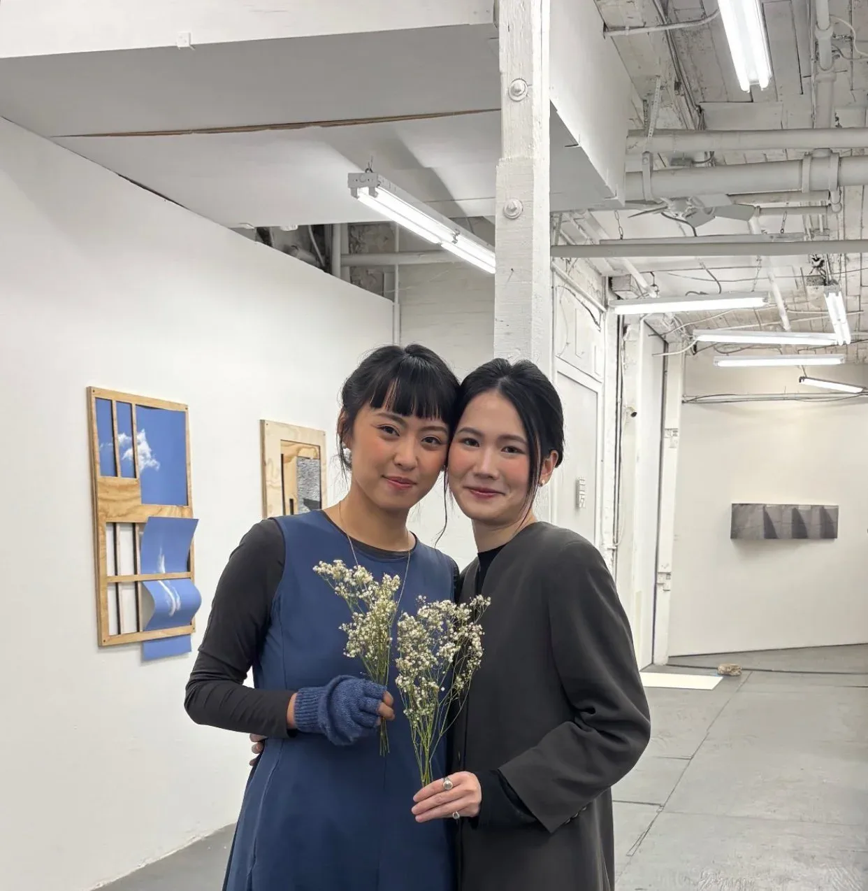 Two young women posing together in gallery space, one wearing navy dress, holding white flowering plant.