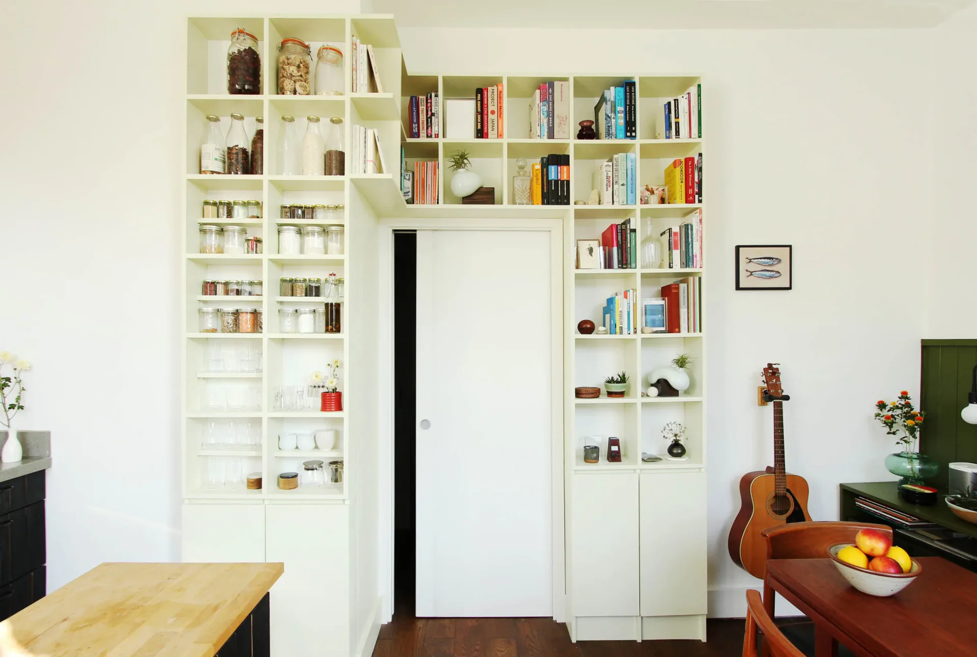 Residential interior with extensive modular white shelving surrounding a dark doorway, displaying books and objects; wooden table in foreground, violin visible on right.