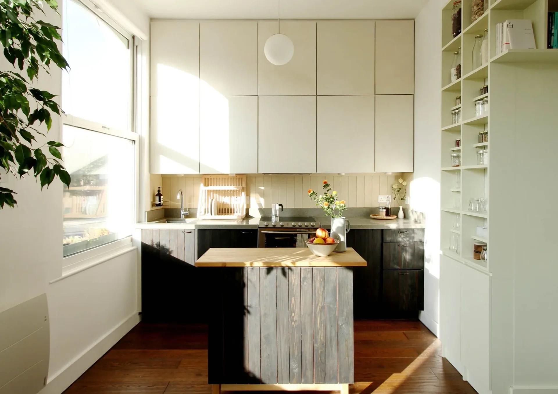 Compact kitchen interior with cream upper cabinets, black lower cabinets, natural wood island, open shelving, and bright window with morning light.