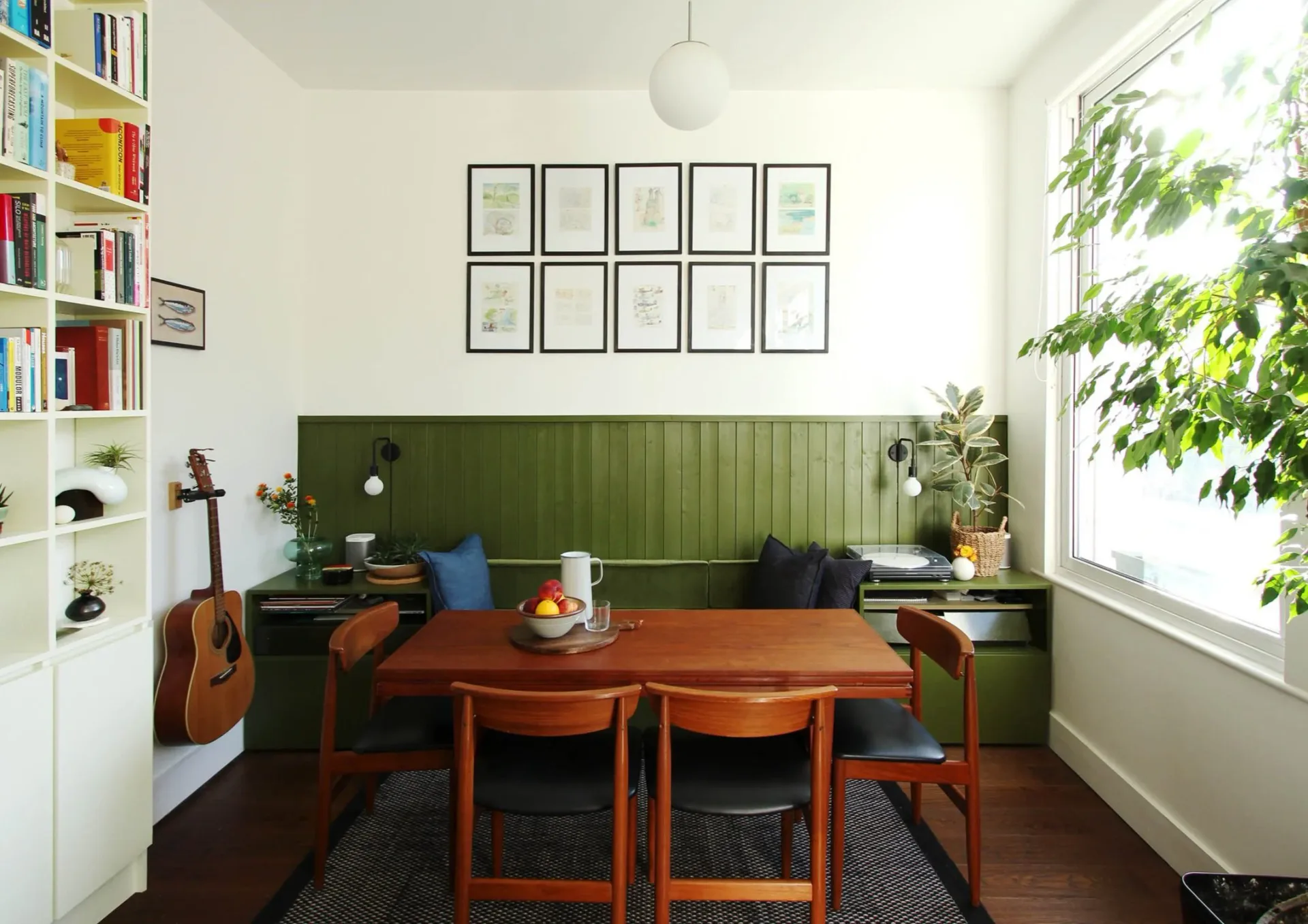 Dining room with green vertical shiplap accent wall, wooden table with mid-century chairs, white shelving, grid of framed prints, violin and potted plants visible.