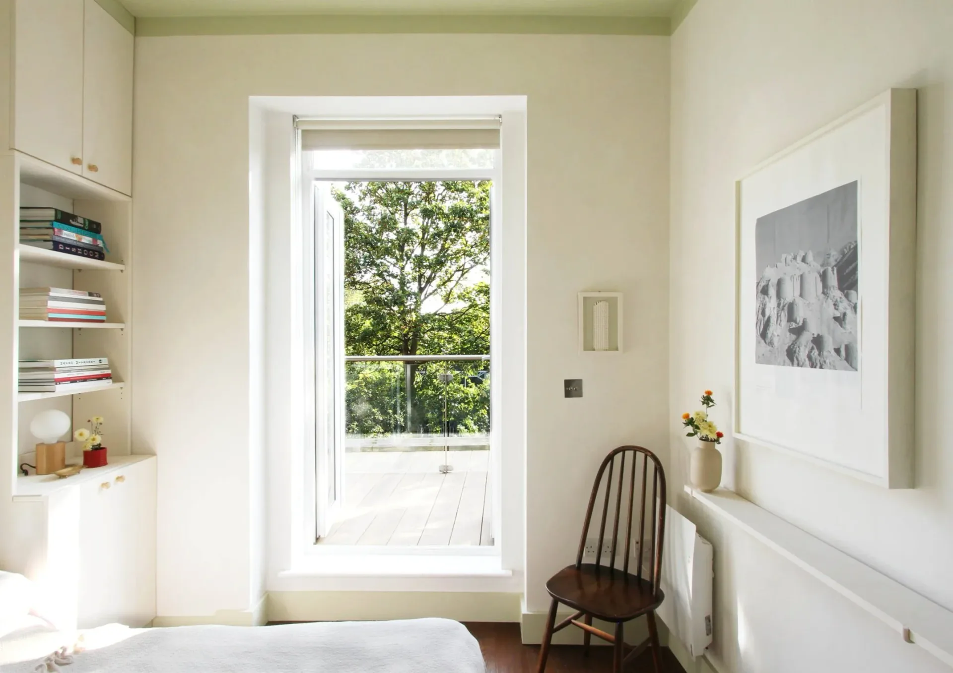 Bright bedroom or office nook with white built-in shelving on left, tall multi-pane window showing green trees, dark wooden chair, and neutral cream walls.