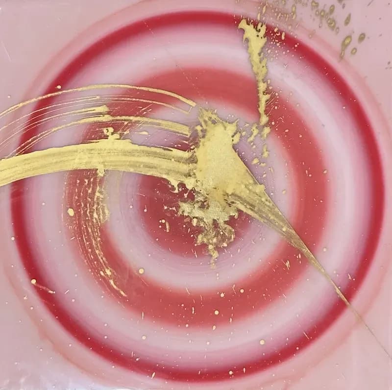 Paintbrush with yellow pigment swirling in circular pink and red liquid base, creating dynamic abstract pattern.